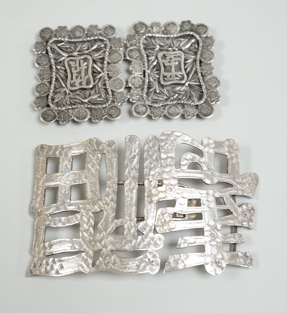 A Chinese white metal belt buckle, 13.3cm wide, the border cast with models of Guangxu cash and a Chinese plated ‘fu Shou’ belt buckle